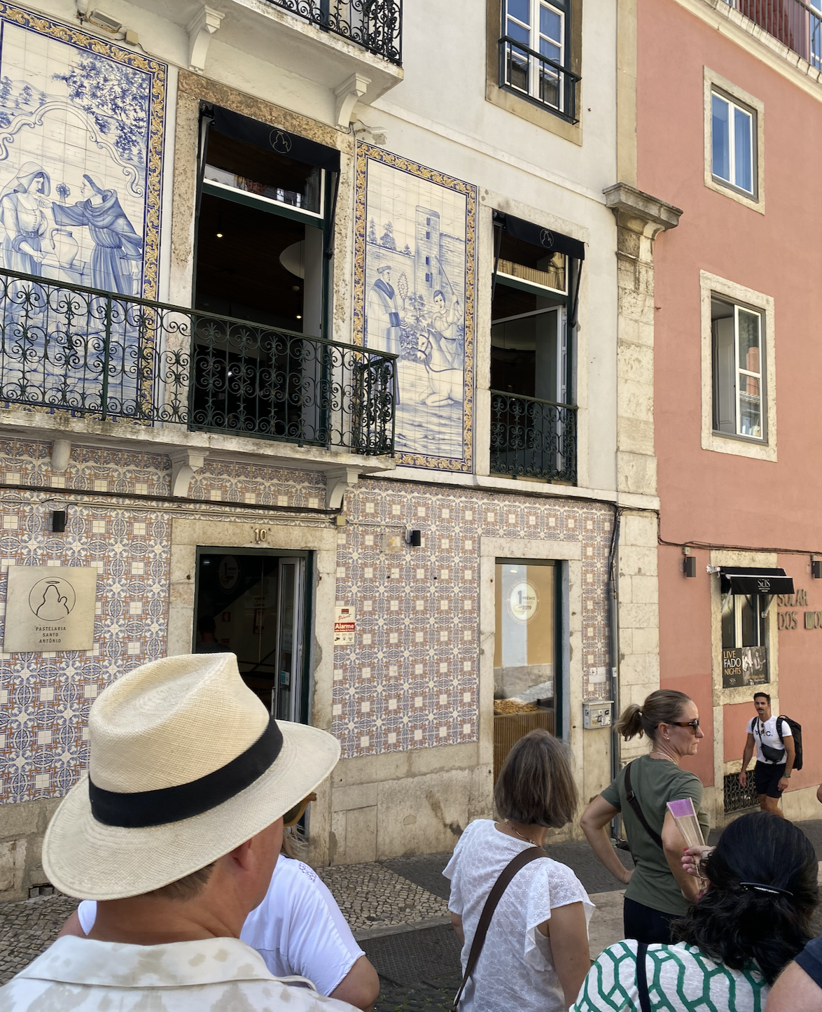 Turister står foran farverige bygninger med azulejo-mosaikker, åbne vinduer og en stor hat i forgrunden.
